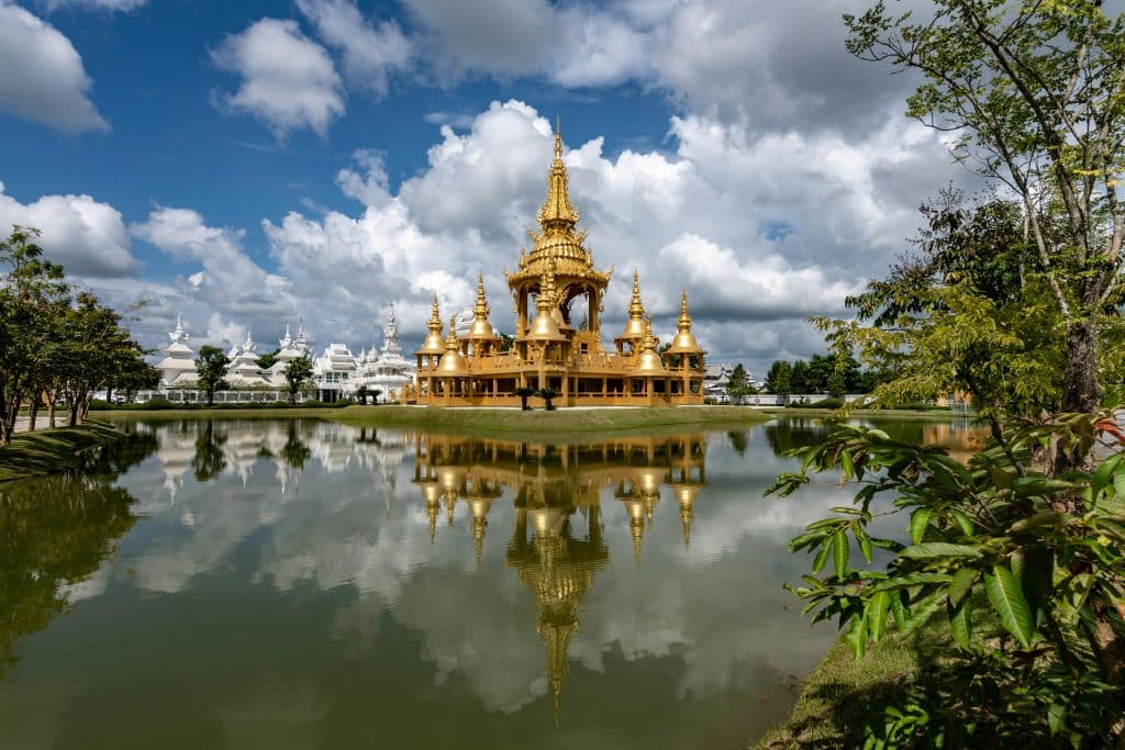 Wat Rong Khun i Chiang Rai, Thailand – ikoniskt vitt tempel och gyllene byggnad vid spegelblank sjö under dramatisk himmel