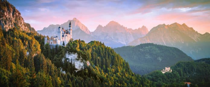 Neuschwanstein slott i Bayern omgitt av de tyske Alpene ved solnedgang, med dramatiske fjell og frodig skog i bakgrunnen