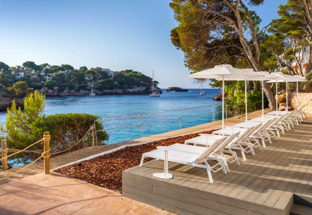 Här är ett passande alt-tekstförslag på svensk: Havsområde i Cala d’Or med kristallklart turkost vatten, små vikar och vitkalkade byggnader längs kusten i soligt medelhavsklimat.