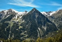 Bergslandskap i Andorra i Pyrenéerna med snötäckta toppar, slingrande bergsväg och grön dal under klarblå himmel