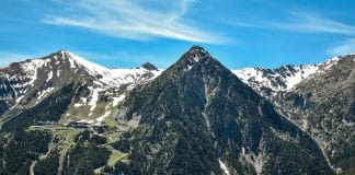 Bergslandskap i Andorra i Pyrenéerna med snötäckta toppar, slingrande bergsväg och grön dal under klarblå himmel