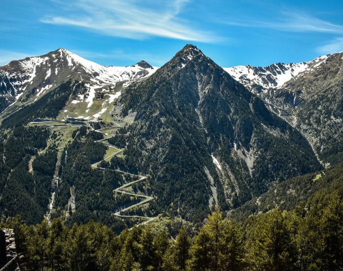 Bergslandskap i Andorra i Pyrenéerna med snötäckta toppar, slingrande bergsväg och grön dal under klarblå himmel