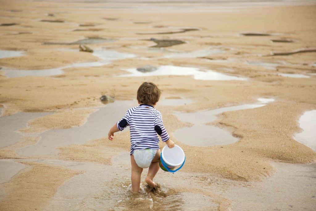 Litet barn som leker i vattenbrynet på en sandstrand med hink i handen – en stillsam och barnvänlig strandupplevelse som passar perfekt för familjer som reser med små barn.