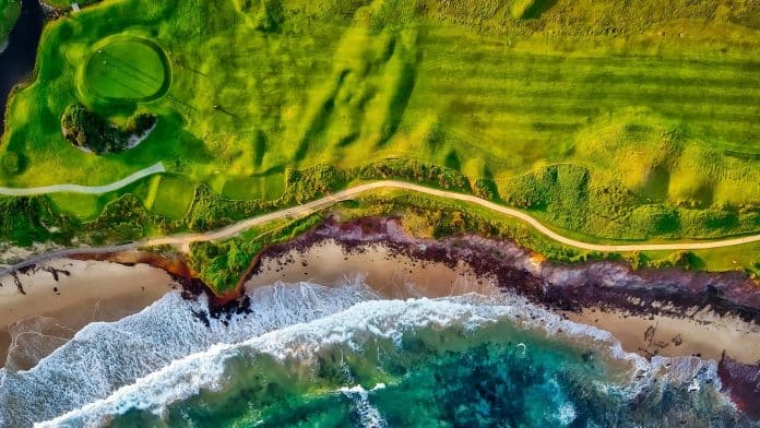 Lyxig golfbana vid havet med gröna fairways, sandstrand och turkost vatten – flygfoto över kustnära golfresort i Spanien